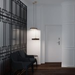 Lampa wisząca Artdeco 3xE14 w kolorze złotego szczotkowanego i czarnym, z dwoma okrągłymi kloszami. Zawieszona na cienkich linkach, w otoczeniu nowoczesnego wnętrza z czarnymi fotelami i dekoracyjną ścianką.