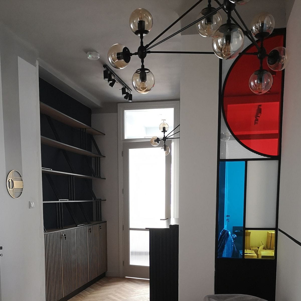 Lampa wisząca Astrifero, czarna z bursztynowymi kloszami, składająca się z 21 opraw. Widoczna w nowoczesnym wnętrzu, zawieszona na cienkich metalowych ramionach. W tle półki oraz kolorowe witraże w odcieniach czerwieni i niebieskiego.