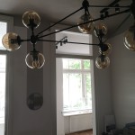 Lampa wisząca Astrifero w kolorze czarnym z bursztynowymi kloszami, z 21 miejscami na żarówki E27. Umieszczona w centralnej części pomieszczenia, z widokiem na okno i jasne tło. Klosze mają okrągły kształt, a całość utrzymana w nowoczesnym stylu.