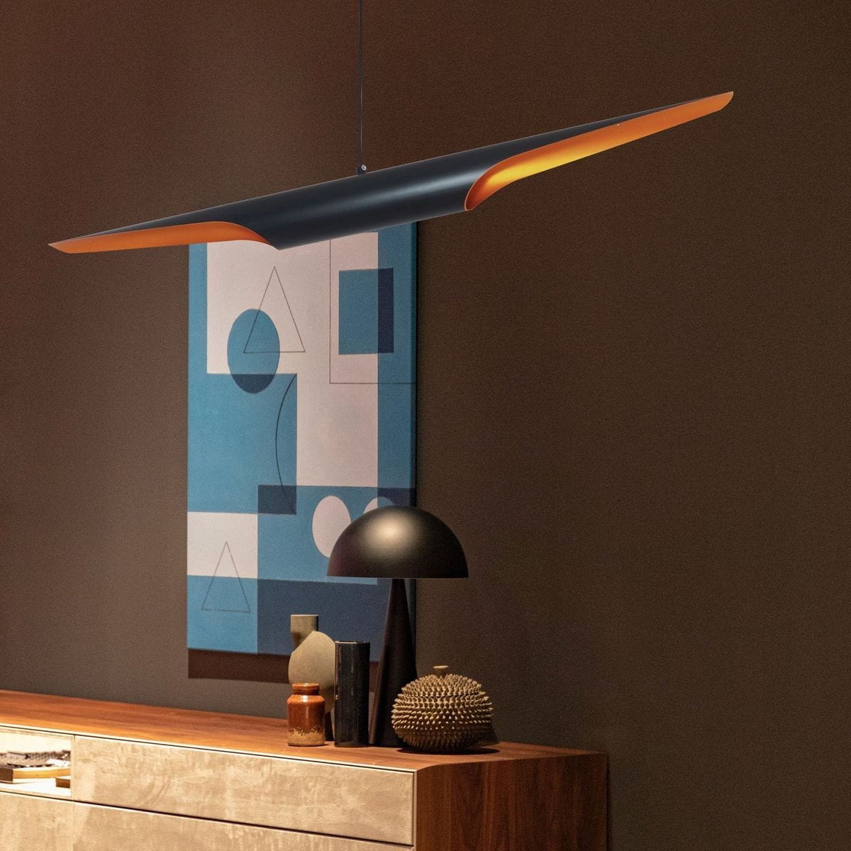 Lampa wisząca Black Tube 2xE27 w kolorze czarnym z złotym wykończeniem, zawieszona na cienkim przewodzie. Widok z przodu, z tłem w odcieniach brązu i niebieskiego, obok drewnianego mebla oraz dekoracji.