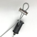 Zbliżenie na element mocujący do lampy wiszącej Black Tube. Widać szary przewód oraz metalowy klips, który trzyma przewód w miejscu. Element jest czarny, wykonany z tworzywa, z widocznym mechanizmem mocującym. Tło jasne, neutralne.