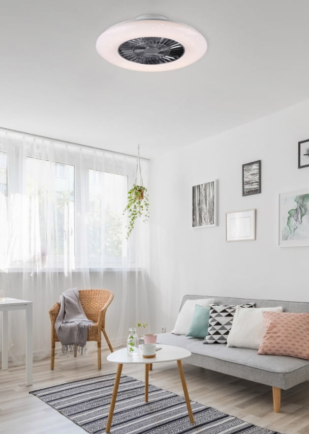 Lampa sufitowa Visby z wentylatorem w kolorze białym i chromowanym, z pilotem, umieszczona na suficie w jasnym wnętrzu. Okrągła konstrukcja z wbudowanym wentylatorem i LED 38W. Otoczenie: przestronny pokój dzienny z kanapą, stolikiem i roślinnością.