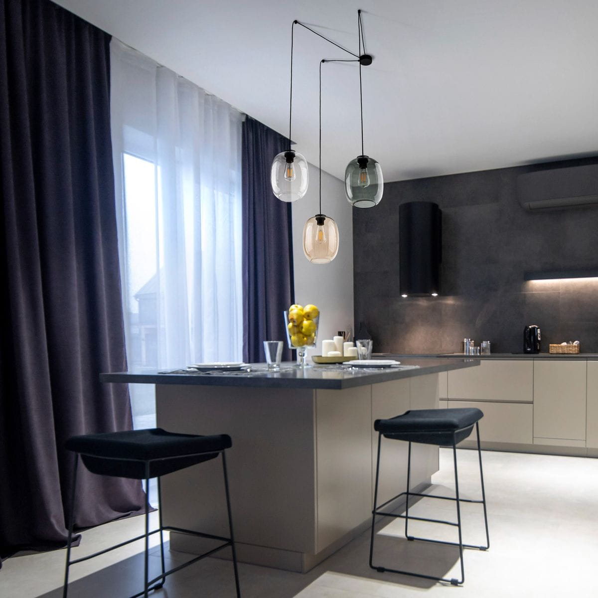 Lampa wisząca Elio z trzema żarówkami E27 w kolorze czarnym i multikolorowym, zawieszona nad blatem kuchennym. Szklane klosze w różnych kolorach, w nowoczesnym wnętrzu z ciemnymi zasłonami i jasnymi meblami.