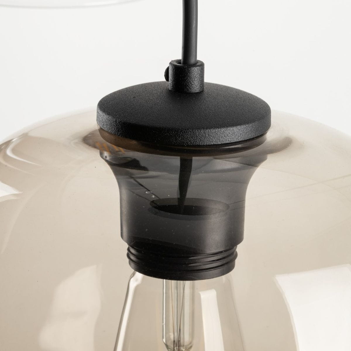 Lampa wisząca Elio z trzema gwintami E27, czarna podstawa z matowego metalu, przezroczysta klosz w odcieniu brązowym. Ujęcie z bliska, pokazujące detal mocowania i przewodu.