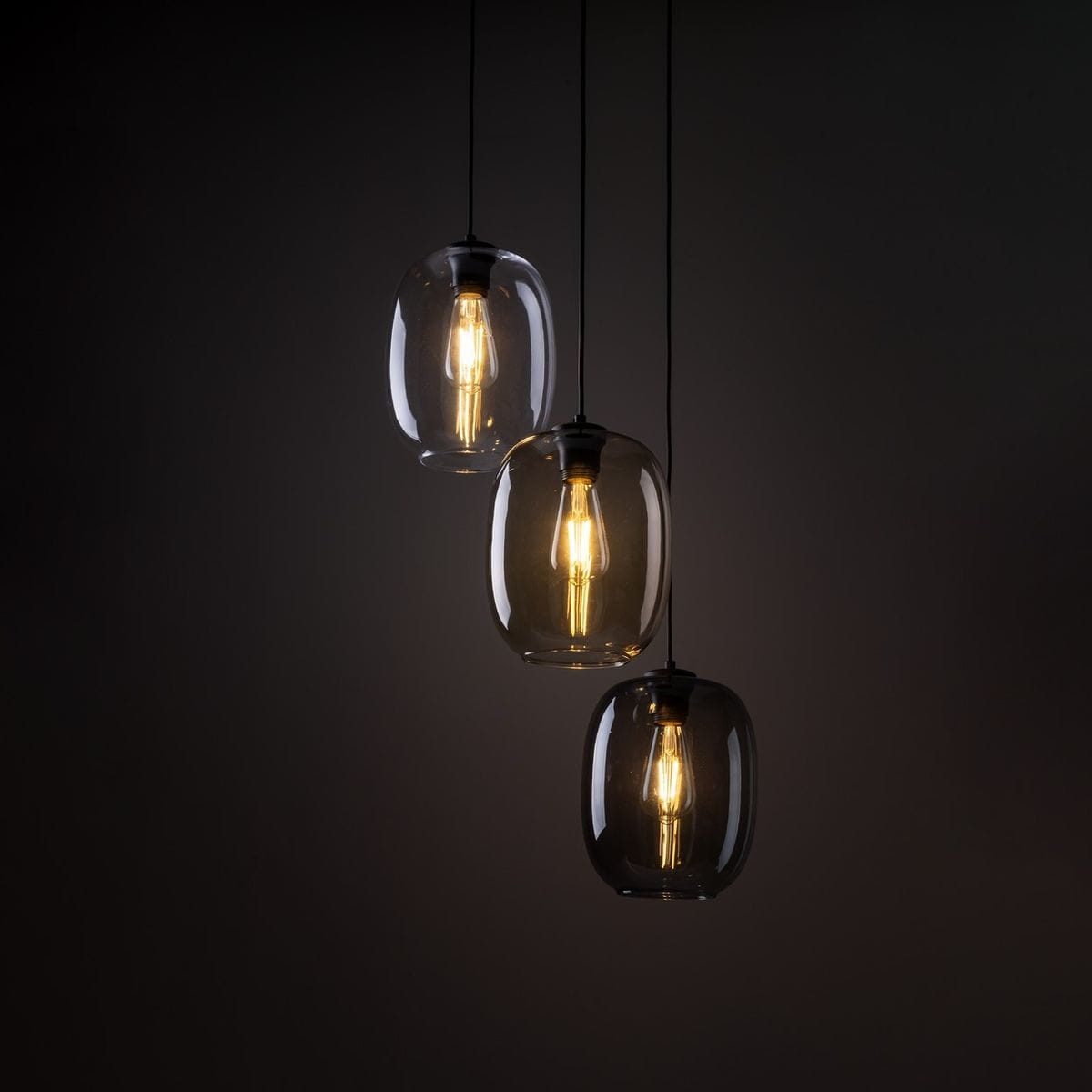 Lampa wisząca Elio z trzema kloszami w kształcie owalnym, wykonanymi z ciemnego szkła, z żarówkami E27. Zawieszona na czarnych przewodach, idealna do nowoczesnych wnętrz. Ujęcie z przodu, na ciemnym tle.