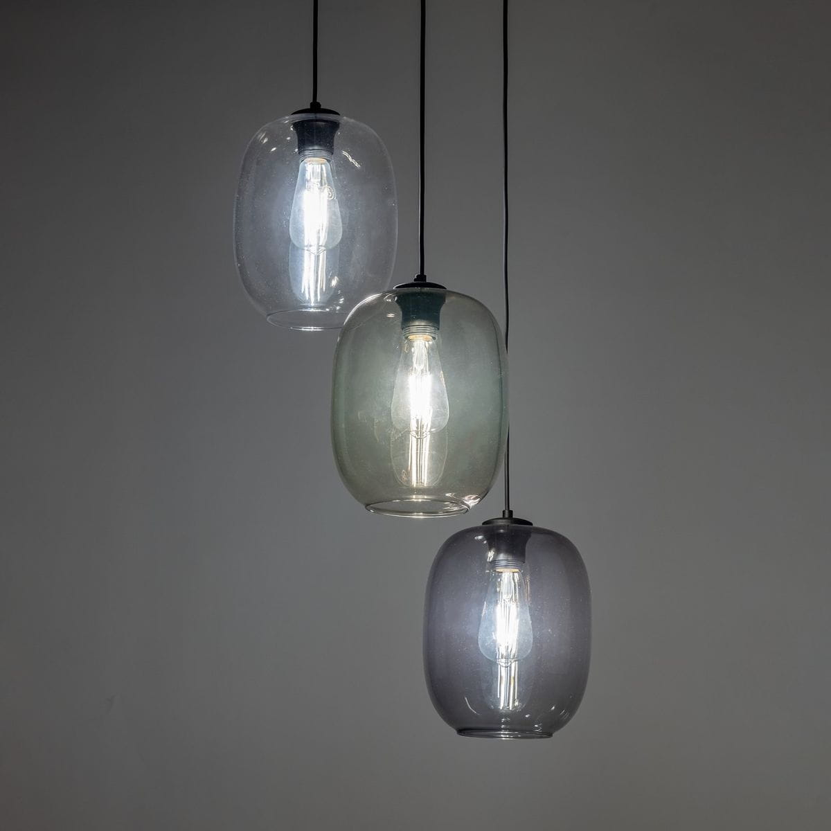 Lampa wisząca Elio z trzema kloszami w różnych odcieniach szarości, wykonana z przezroczystego szkła. Klosze zawieszone na czarnych przewodach, z widocznymi żarówkami E27. Tło ciemnoszare, minimalistyczne.