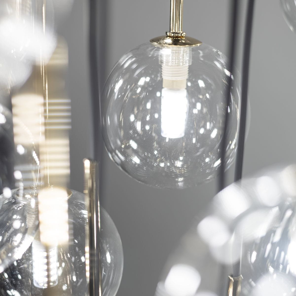 Lampa wisząca Aurora z 16 żarówkami G9 w czarnej i przezroczystej obudowie. Widoczne szklane klosze w kształcie kul, metalowe elementy w kolorze złotym, zawieszone na cienkich przewodach. Tło w odcieniach szarości.