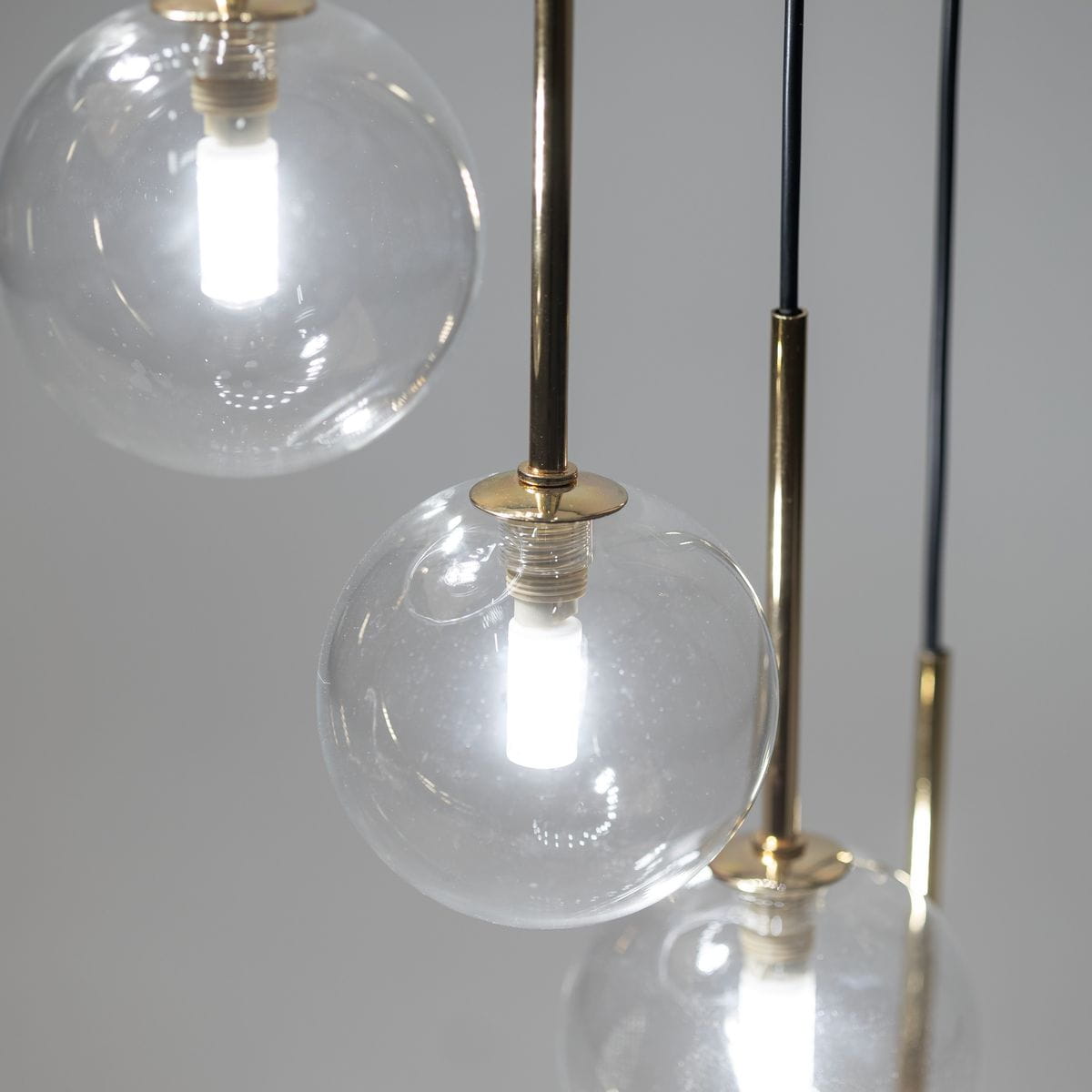 Lampa wisząca Aurora z 16 źródłami światła G9, w kolorze czarnym i transparentnym. Zbliżenie na trzy szklane kule, w których widoczne są źródła światła, zawieszone na złotych metalowych stelażach. Tło w odcieniu szaro-białym.