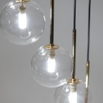 Lampa wisząca Aurora z 16 źródłami światła G9, w kolorze czarnym i transparentnym. Zbliżenie na trzy szklane kule, w których widoczne są źródła światła, zawieszone na złotych metalowych stelażach. Tło w odcieniu szaro-białym.