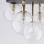 Lampa sufitowa Nilos z pięcioma kloszami w kształcie kul, wykonana z transparentnego szkła. Elementy montażowe w kolorze czarnym z złotymi akcentami. Widoczne źródła światła G9 w przezroczystych kloszach. Ujęcie z bliska, pod kątem.