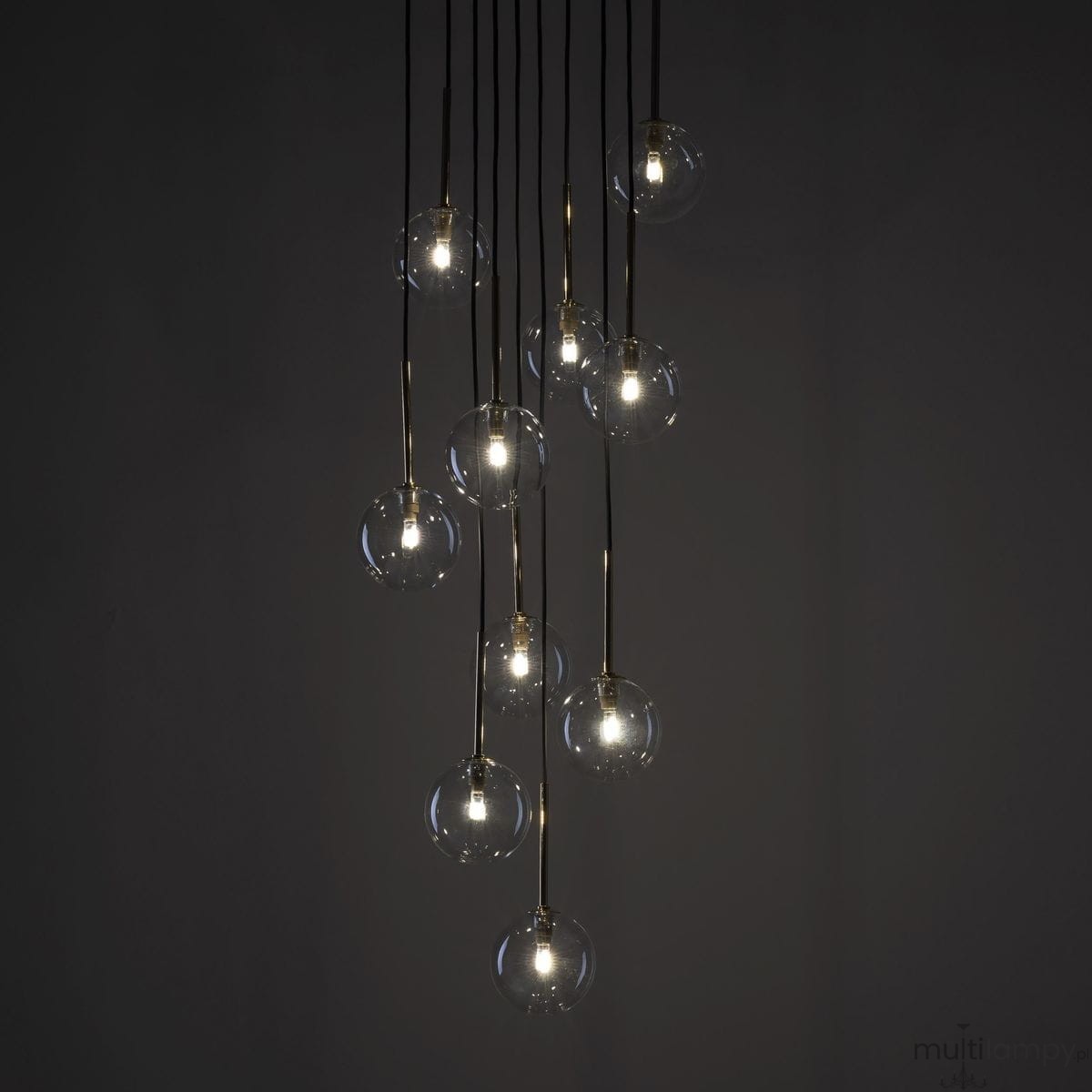 Lampa wisząca Nilos z 10 żarówkami G9, w czarnej i transparentnej kolorystyce. Składa się z okrągłych, szklanych kloszy zawieszonych na cienkich, złotych linkach. Ujęcie z przodu, prezentujące efektowne oświetlenie w ciemnym tle.