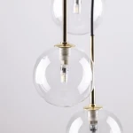 Lampa wisząca Nilos z siedmioma przezroczystymi kloszami w kształcie kul, zawieszona na złotych mocowaniach. Widać gwinty G9, w których umieszczone są źródła światła. Tło jasne, podkreślające elegancki design.