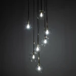 Lampa wisząca Nilos z siedmioma przezroczystymi kloszami, zawieszona na czarnych przewodach. Klosze mają okrągły kształt, a w środku znajdują się żarówki G9. Całość prezentuje nowoczesny design na ciemnym tle.