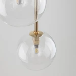 Lampa wisząca Nilos z trzema kloszami G9 w kolorze czarnym i transparentnym. Klosze w kształcie kul, wykonane ze szkła, z widocznymi żarówkami LED i złotym mocowaniem. Ujęcie z góry, ukazujące detale konstrukcji.