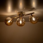 Lampa sufitowa Estera z trzema kulistymi kloszami w kolorze bursztynowym, zamontowana na złotej metalowej podstawie. Ujęcie z dołu, ukazujące źródła światła wewnątrz kloszy. Klosze mają przezroczysty wygląd, emitując ciepłe światło.
