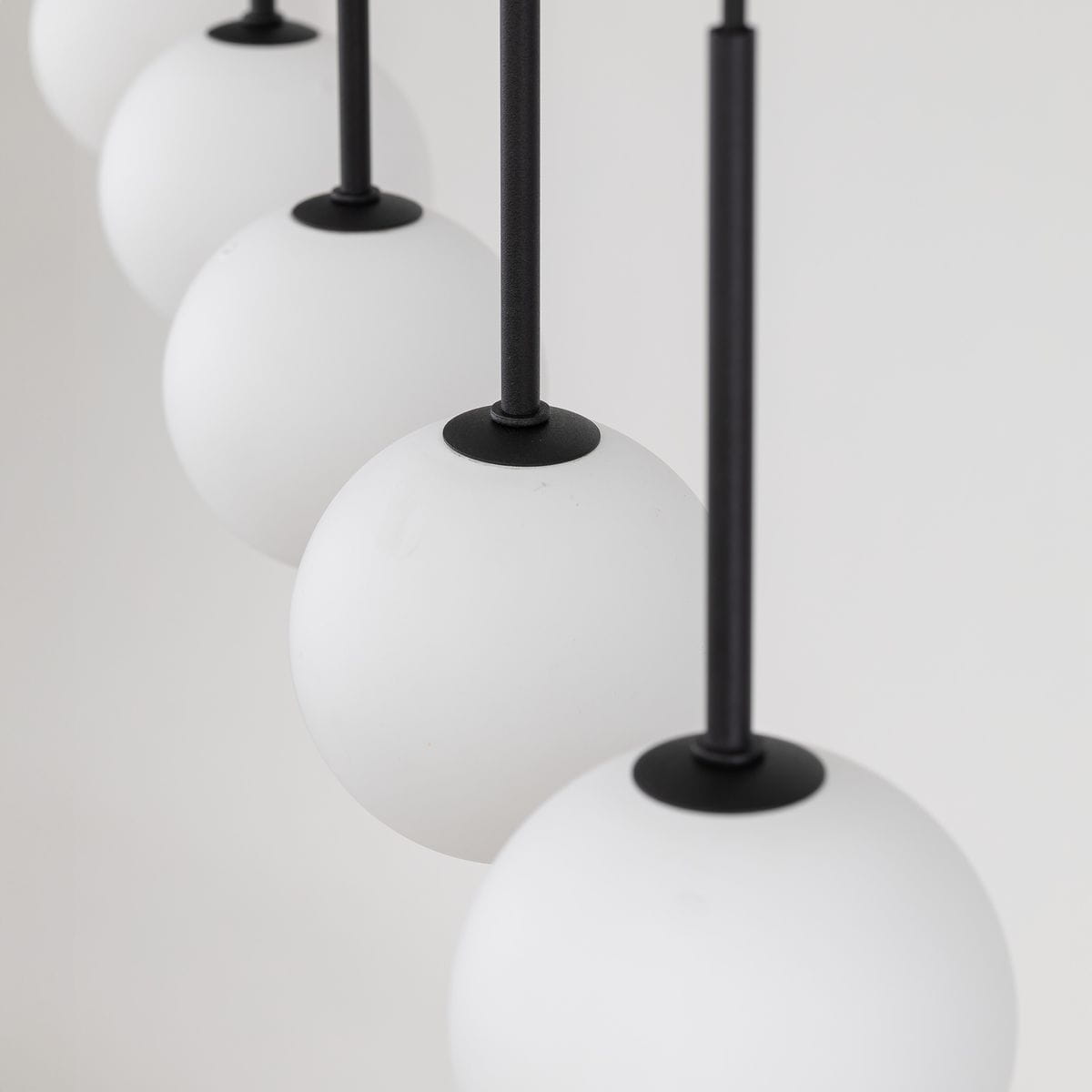 Lampa wisząca Martin 6xG9 w kolorze czarnym. Składa się z sześciu białych, okrągłych kloszy z matowego szkła zawieszonych na cienkich, czarnych prętach. Zbliżenie na detale mocowania i kloszy, na jasnym tle.