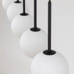 Lampa wisząca Martin 6xG9 w kolorze czarnym. Składa się z sześciu białych, okrągłych kloszy z matowego szkła zawieszonych na cienkich, czarnych prętach. Zbliżenie na detale mocowania i kloszy, na jasnym tle.