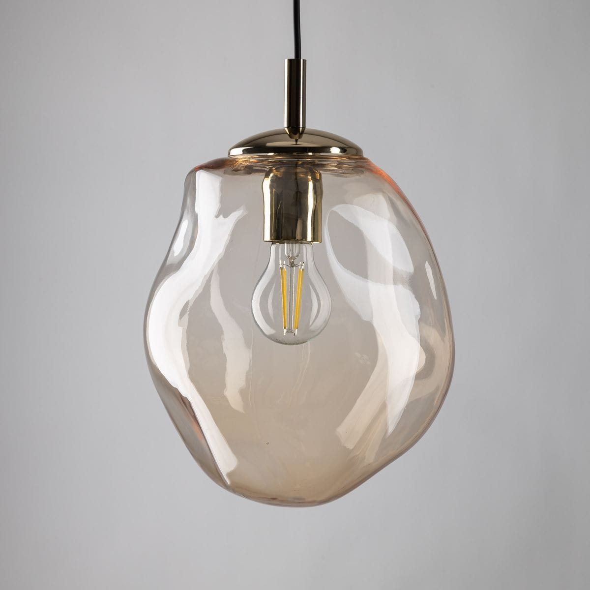 Lampa wisząca Sol D25 z kloszem w bursztynowym kolorze, wykonana z przezroczystego szkła. U podstawy złoty element mocujący z oprawką na żarówkę E27. Produkt zwisający na czarnym przewodzie, prezentujący nowoczesny design.