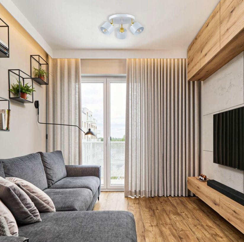 Okrągła lampa sufitowa Elza z trzema oprawami E27, w kolorze białym z złotymi akcentami. Umieszczona na suficie w nowoczesnym wnętrzu, z szarym narożnikiem i drewnianym wykończeniem. Tło z jasnymi zasłonami i dużym oknem.