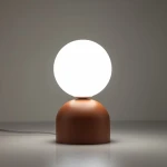 Lampka stołowa Miki z podstawą w kolorze czerwonym i białą, okrągłą żarówką. Ujęcie z boku pokazuje okrągłą formę podstawy oraz gładką powierzchnię. Produkt leży na jasnej powierzchni, w neutralnym otoczeniu.