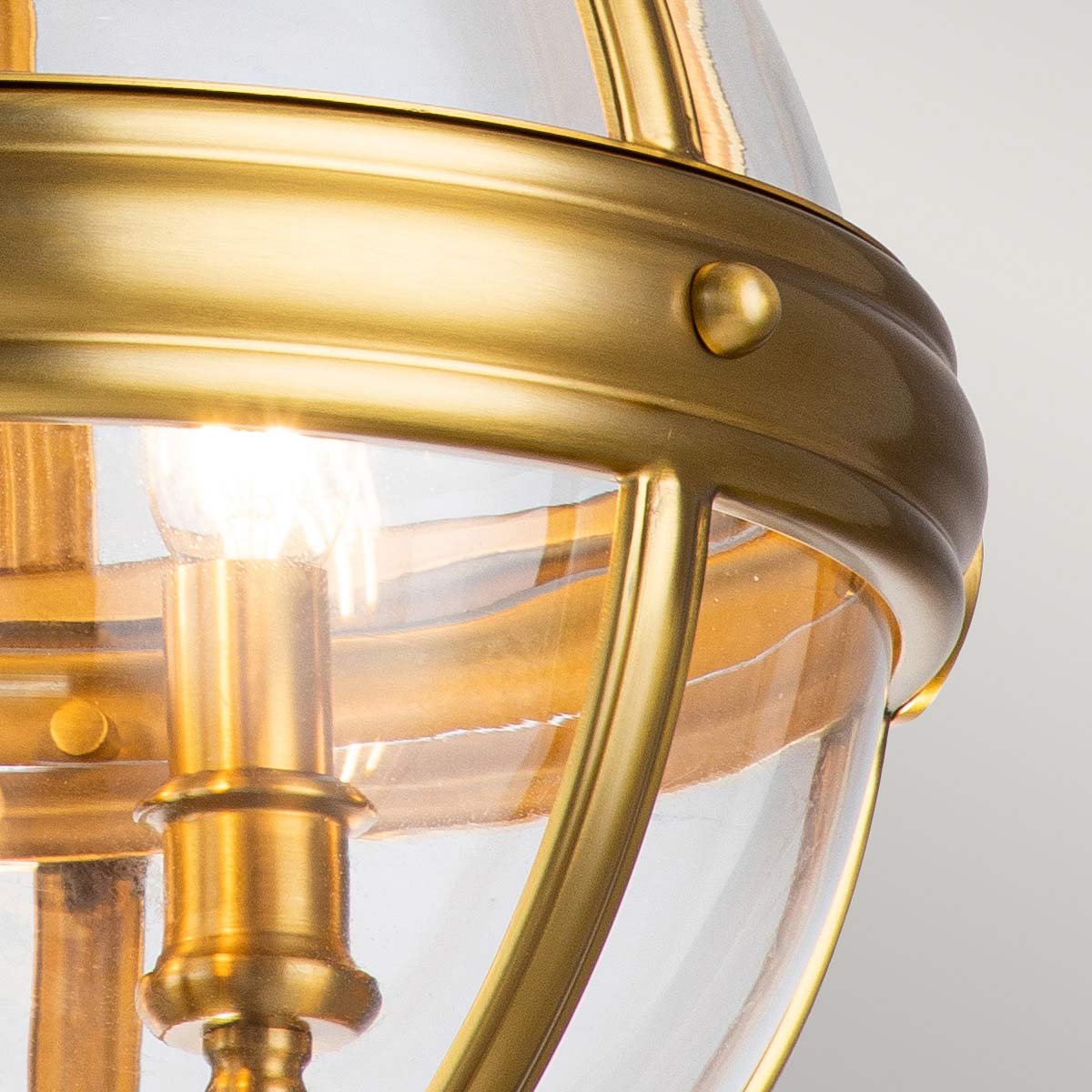 Mosiężna lampa wisząca Adams z trzema oprawkami E14, z przezroczystym kloszem. Widoczny detal metalowej obręczy i żarówki w centralnej części, z bliska, na jednolitym tle.