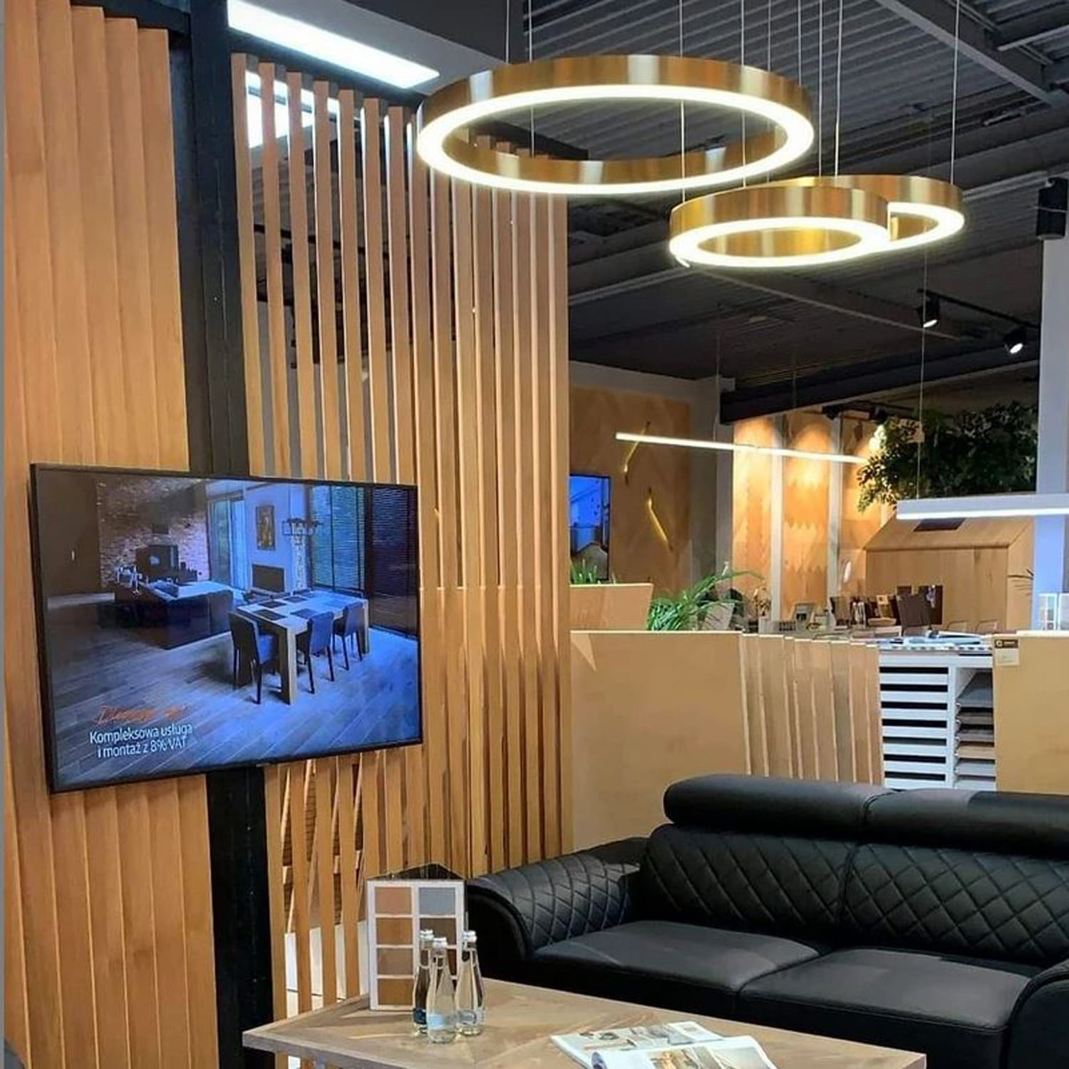 Lampa wisząca Circle 40+60+80 w kolorze mosiądzu, z trzema okrągłymi pierścieniami LED, zawieszona nad czarną sofą w nowoczesnym wnętrzu. Tło z drewnianymi panelami i telewizorem. W zestawie pilot do sterowania.