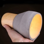 Lampa wisząca Concrete z szarym betonem i drewnianym uchwytem, z otworem do żarówki E27. Ujęcie z ręką trzymającą lampę, pokazujące matową teksturę betonu i ciepłe, żółte światło wewnątrz klosza.