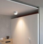 Lampa sufitowa Grandi w kształcie tuby, biała, z oprawą 1xGX53, zamontowana na suficie. Emituje ciepłe światło, umiejscowiona nad białą szafą w minimalistycznym wnętrzu. Tło w odcieniach szarości.
