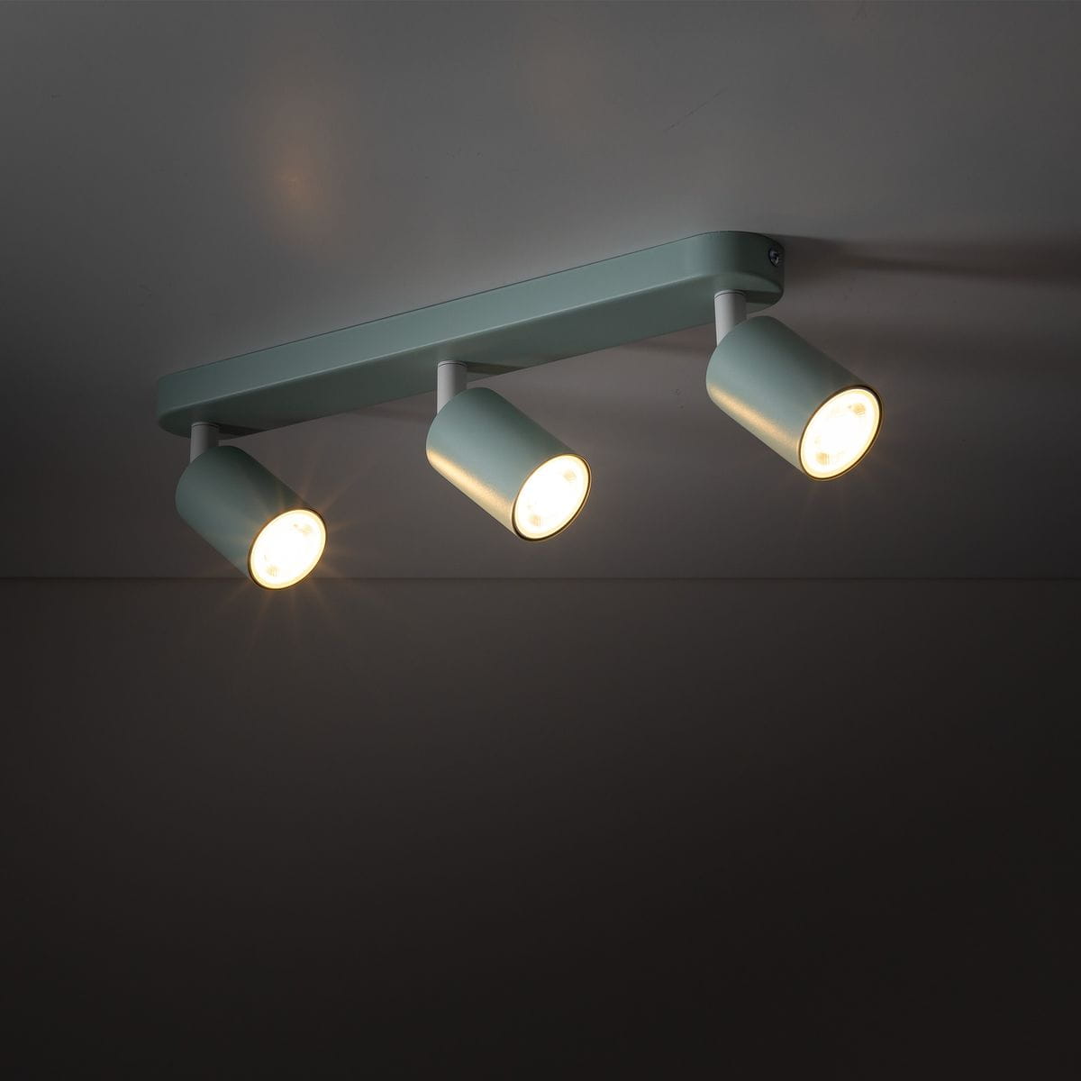Miętowa lampa sufitowa Livia z trzema reflektorami GU10, zamontowana na prostokątnej podstawie. Reflektory mają cylindryczny kształt i są ustawione w różnych kierunkach, emitując jasne światło. Tło ciemne, skupione na produkcie.
