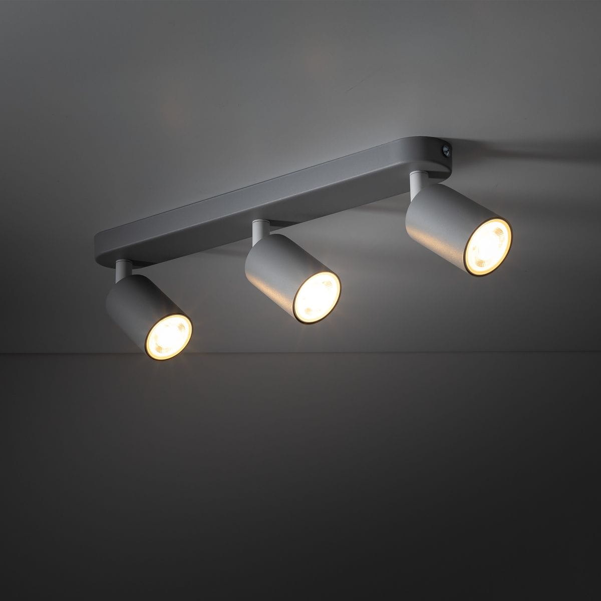 Beżowa lampa sufitowa Livia z trzema spotami na żarówki GU10. Ujęcie z góry, ukazujące zamontowane oprawy o cylindrycznym kształcie, emitujące ciepłe światło, zamocowane na prostokątnej podstawie. Tło ciemne, co podkreśla detal lampy.