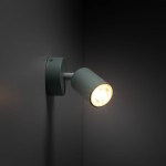 Kinkiet Livia w kolorze miętowym, z metalowym ramieniem i okrągłą podstawą, zamontowany na ścianie. Lampa z żarówką GU10, skierowana w dół, z widocznym źródłem światła. Tło ciemne, podkreślające kolor lampy.