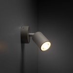 Beżowy kinkiet Livia z jednym punktem świetlnym (GU10), zamontowany na ścianie. Lampa ma cylindryczny kształt klosza oraz metalową podstawę. Ujęcie z boku, ukazujące skierowane w dół światło. Tło w ciemnym odcieniu, podkreślające produkt.