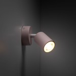 Różowy kinkiet Livia z jedną oprawą GU10, zamontowany na ścianie. Okrągła baza z matowego metalu oraz cylindryczna czasza skierowana w dół, emitująca ciepłe światło. Kąt ujęcia z boku, podkreślający detal konstrukcji.