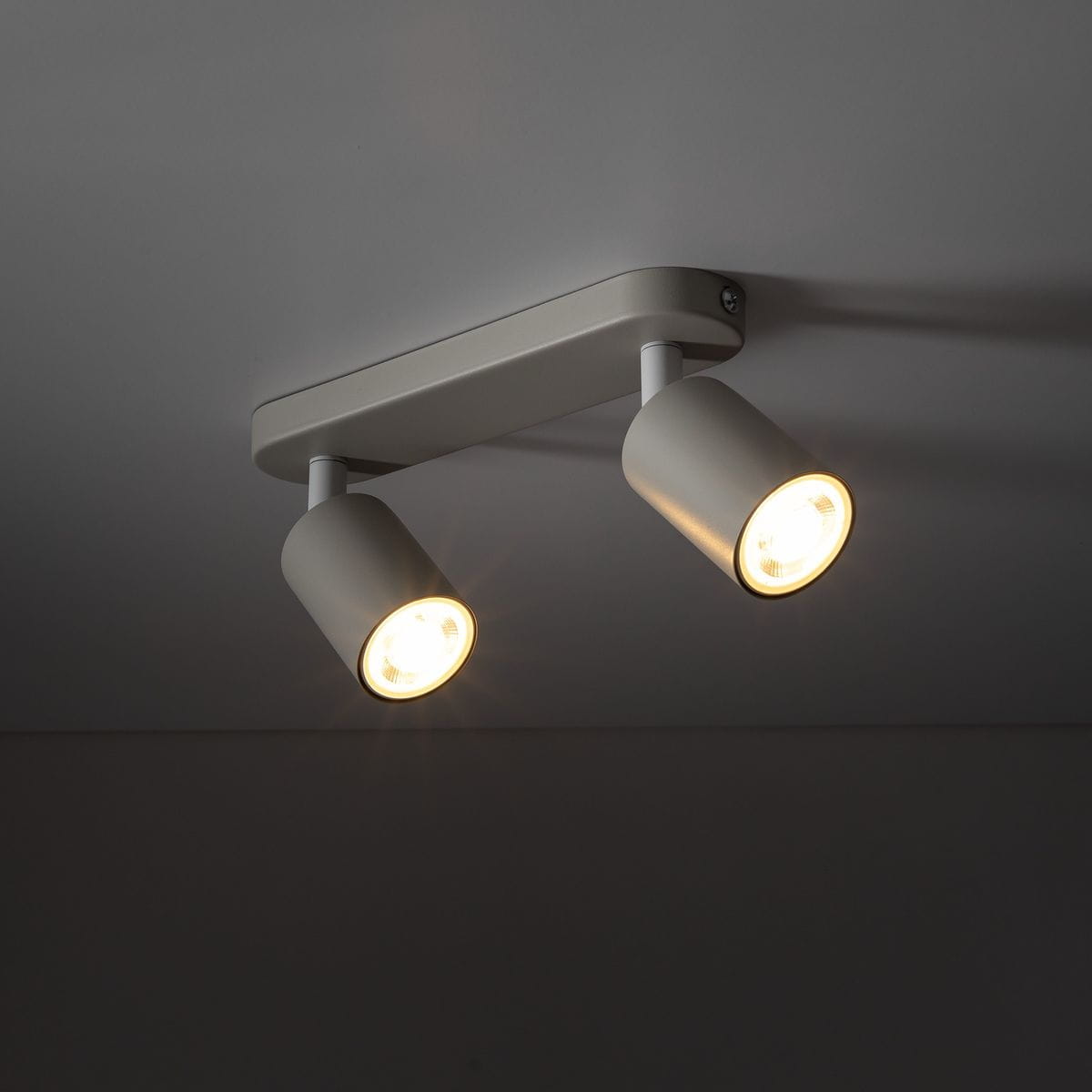 Beżowa lampa sufitowa Livia z dwoma spotami GU10, zamontowana na prostokątnej podstawie. Widoczne dwa cylindryczne klosze, emitujące światło. Ujęcie z dołu, ukazujące detale konstrukcji i oświetlenia.