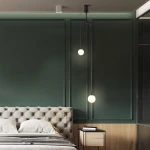 Lampa wisząca Loop 2xG9 w kolorze czarnym, zawieszona na długim przewodzie. Posiada dwie okrągłe, białe żarówki. Zainstalowana w stylowym wnętrzu sypialni, nad łóżkiem i obok nocnej szafki. Tło w ciemnej zieleni podkreśla nowoczesny design.