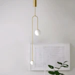 Lampa wisząca Loop 2xG9 w kolorze złotym, wykonana z metalu, z dwiema kulistymi kloszami w kolorze białym, zawieszona na cienkich linkach. Kąt ujęcia z boku, prezentujący minimalistyczny design w jasnym wnętrzu.