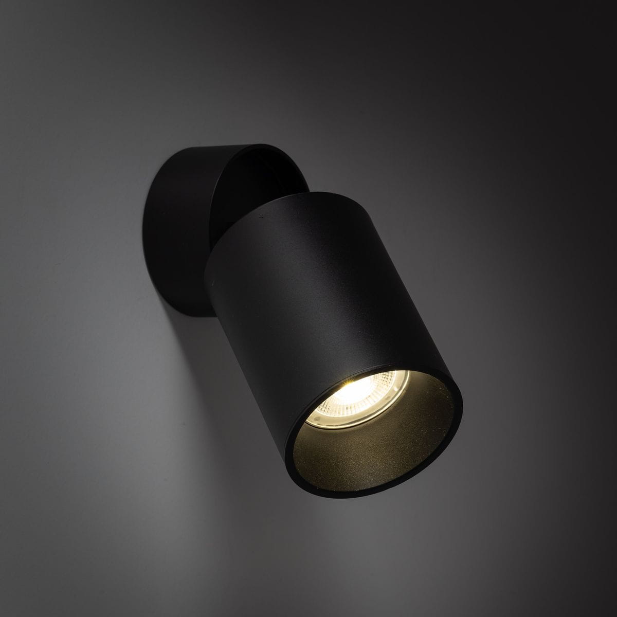Czarna lampa sufitowa typu spot z pojedynczym gwintem GU10, zamontowana na ścianie. Cylindryczny kształt, matowa powierzchnia, skierowana w dół, z widocznym źródłem światła wewnątrz. Tło w odcieniach szarości.