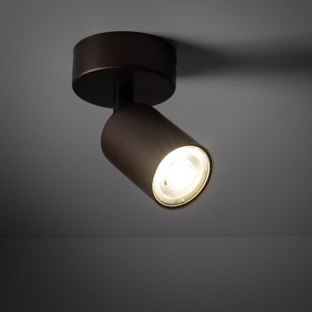 Brązowa lampa sufitowa typu spot z pojedynczym źródłem światła GU10, zamontowana na okrągłej podstawie. Ujęcie z boku, ukazujące zaokrąglony kształt klosza oraz emitowane jasne światło. Tło w kolorze szarym.