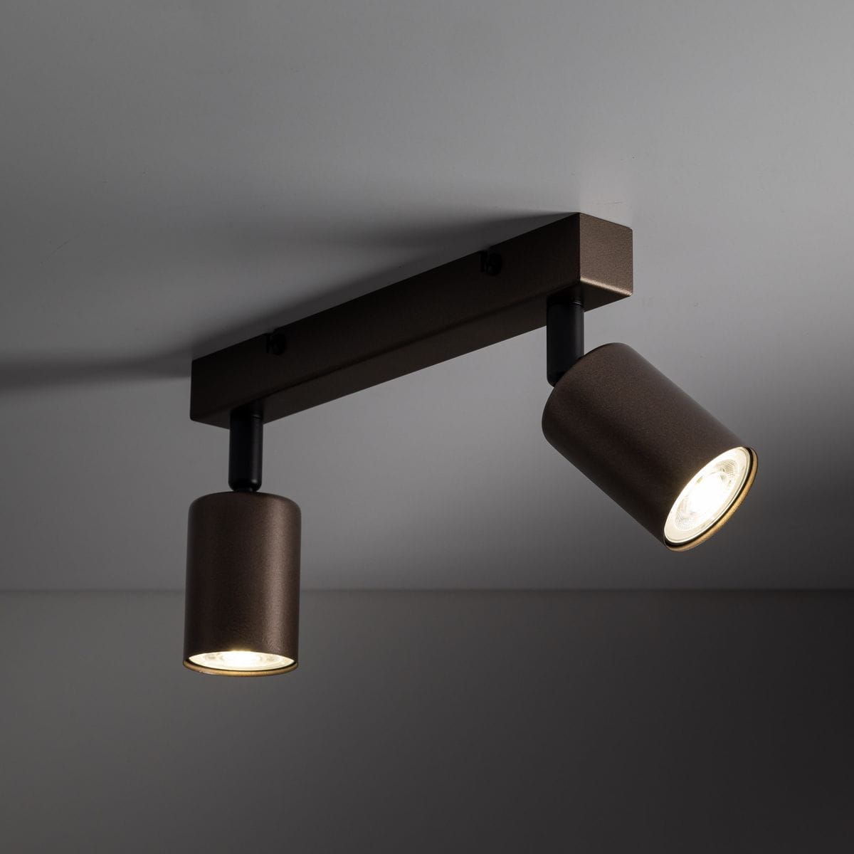 Brązowa lampa sufitowa (spot) z dwoma oprawami na żarówki GU10, zamontowana na prostokątnej, metalowej podstawie. Ujęcie z dołu, pokazujące okrągłe klosze skierowane w różne strony. Tekstura kloszy matowa, z delikatnym połyskiem na krawędziach.