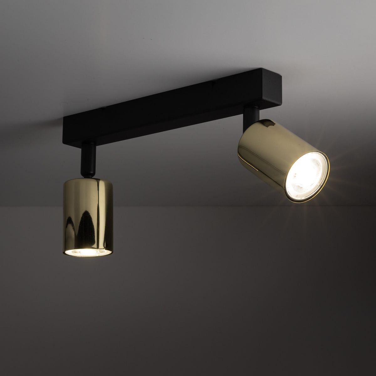 Czarna lampa sufitowa typu spot z dwoma reflektorami, jeden z nich w kolorze złotym. Ujęcie z góry, pokazujące montaż na suficie. Reflektory są cylindryczne i skierowane w różne strony, tworząc efekt oświetlenia.