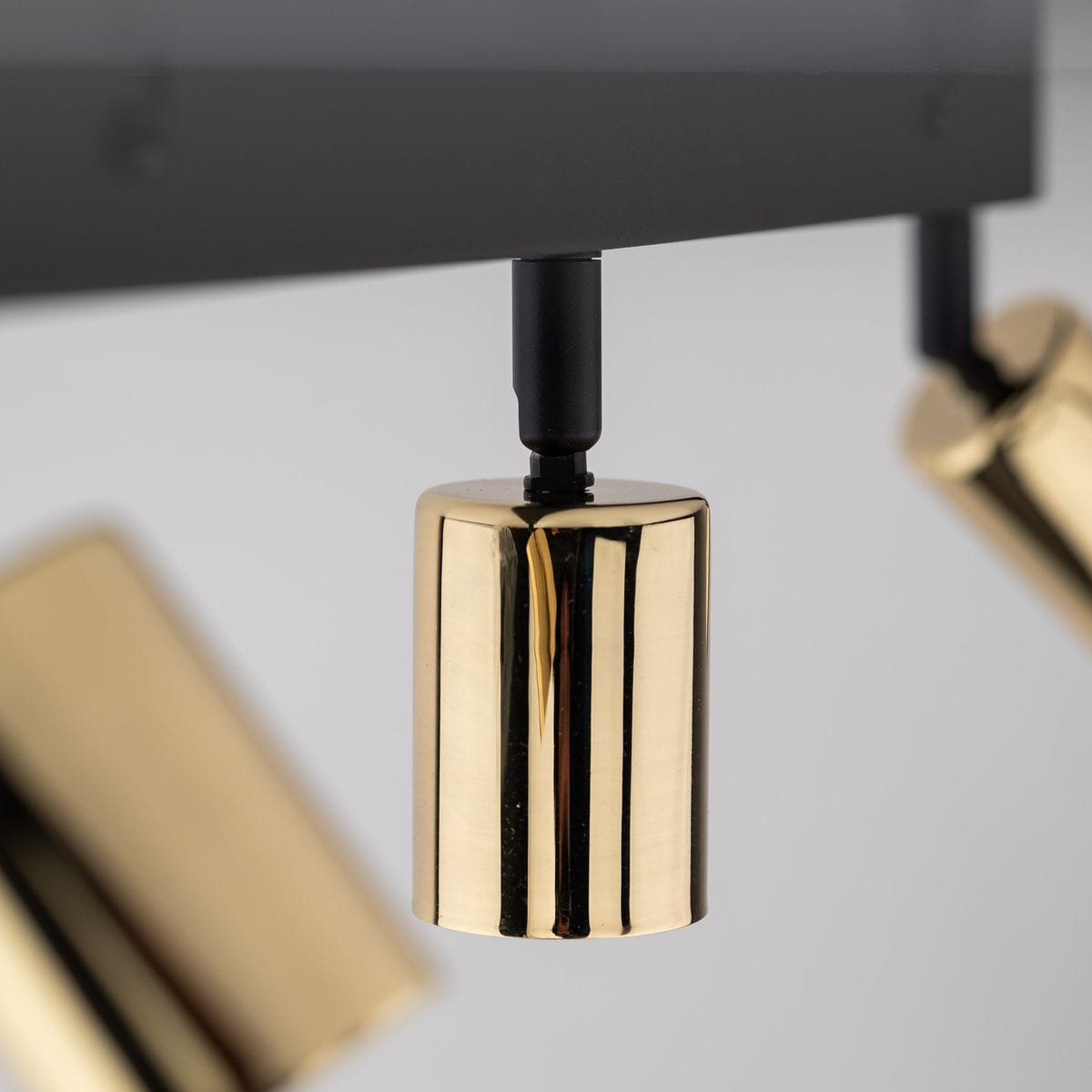 Lampa sufitowa typu spot w kolorze czarnym i złotym z trzema reflektorami na regulowanych uchwytach. Reflektory mają cylindryczny kształt, błyszczącą powierzchnię i są zamontowane na prostokątnej podstawie. Tło jasne.