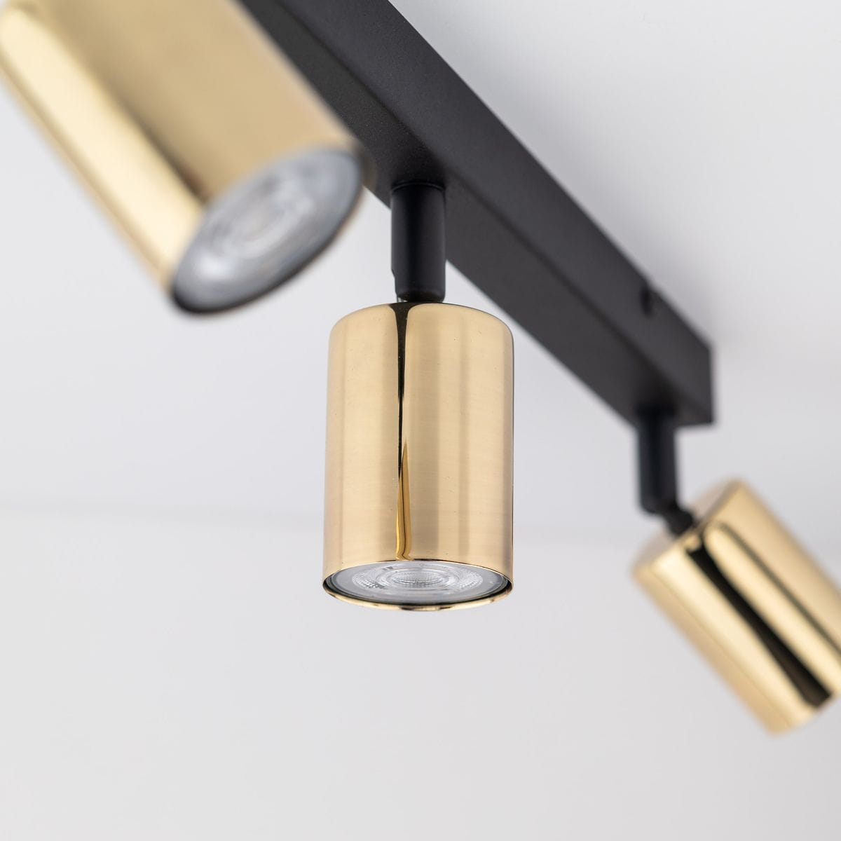 Lampa sufitowa w kolorze czarnym z trzema złotymi reflektorami GU10, zamontowana na prostokątnej podstawie. Reflektory mają cylindryczny kształt, emitując światło w kierunku dołu. Tło jasne, co podkreśla kontrast kolorów.