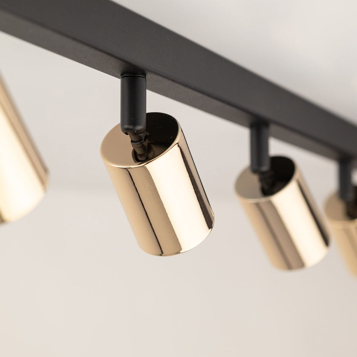 Czarna lampa sufitowa typu spot z ośmioma oprawami GU10. Złote, cylindryczne klosze zawieszone na prostokątnej, czarnej ramie. Ujęcie z bliska, pokazujące detale wykończenia i połączenia. Idealna do nowoczesnych wnętrz.