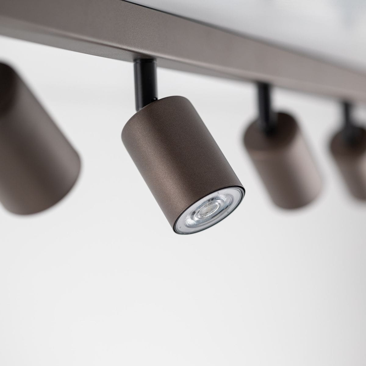Brązowa lampa sufitowa typu spot z ośmioma miejscami na żarówki GU10. Ujęcie z bliska, pokazujące cylindryczne klosze zamontowane na metalowej konstrukcji. Proste tło podkreśla minimalistyczny design lampy.
