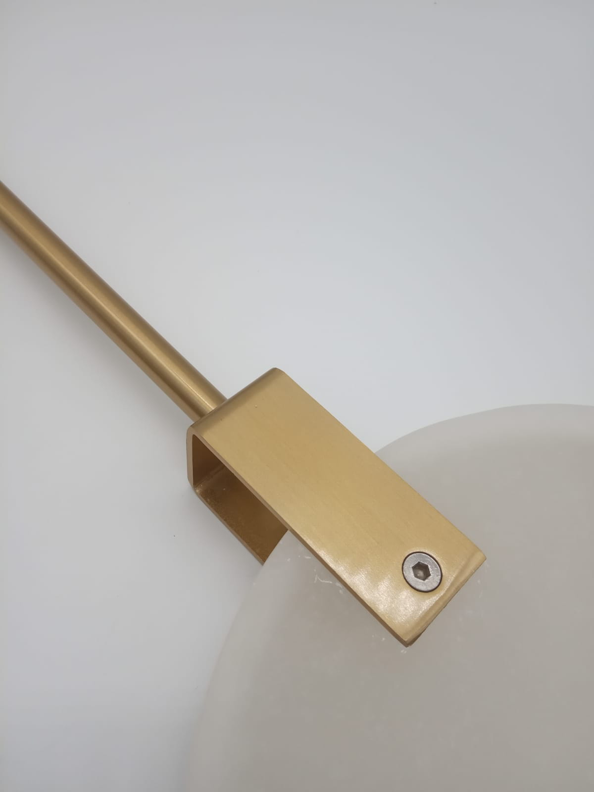 Lampa wisząca Marble D30 z mosiężnym uchwytem, zbliżenie na detal mocowania. Widoczna okrągła, mleczna klosz i prostokątny, złoty element. Tło białe, minimalistyczne, podkreślające elegancję lampy.