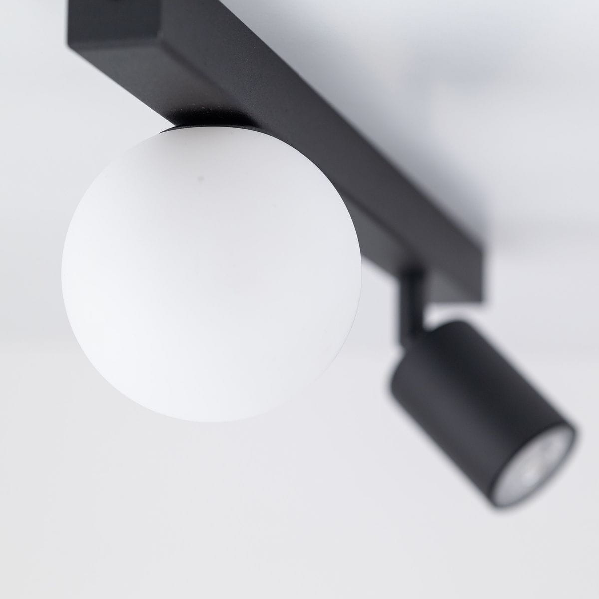 Czarna lampa sufitowa Vox z jednym źródłem światła GU10 i jednym G9. Okrągła, matowa kula w białym kolorze oraz prostokątny, czarny uchwyt. Ujęcie z góry, ukazujące detale konstrukcji.