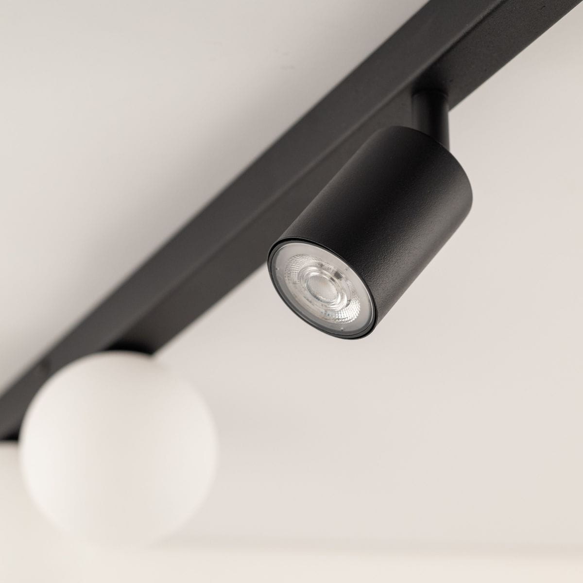 Lampa sufitowa Vox w kolorze czarnym z dwoma spotami GU10 i dwoma kulami G9. Ujęcie pod kątem, pokazujące detal lampy z cylindrycznym reflektorem i białą kulą. Stalowa konstrukcja na jasnym tle.