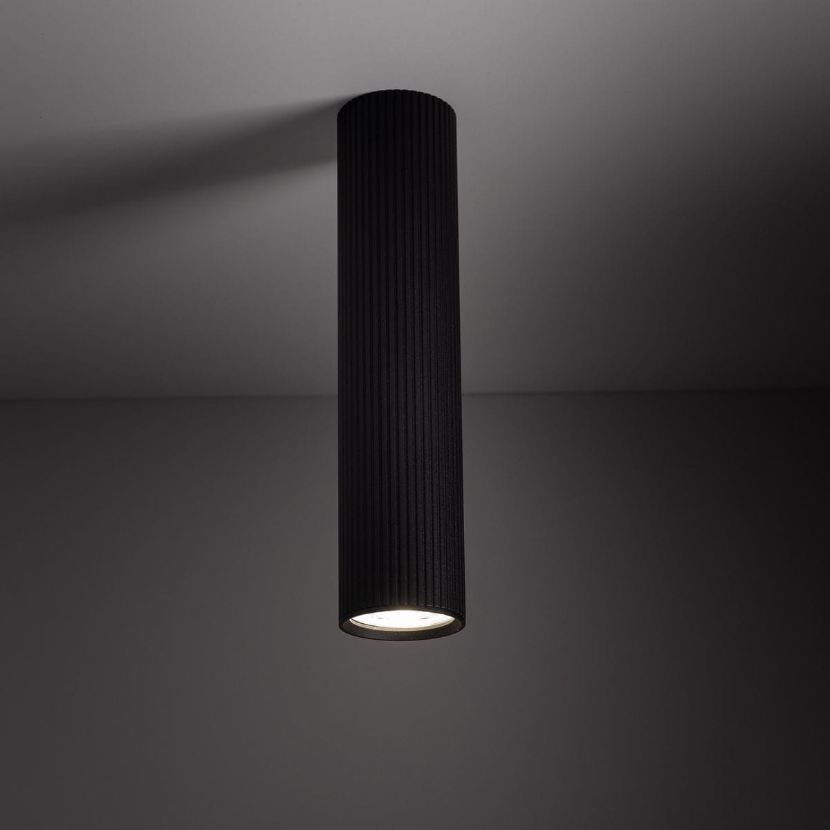 Lampa sufitowa Tune L w kolorze czarnym, cylindryczna, z podłużnymi żłobieniami. Ujęcie z dołu, pokazujące zamontowaną lampę z widocznym źródłem światła GU10. Tło w odcieniach szarości, co podkreśla elegancki design produktu.