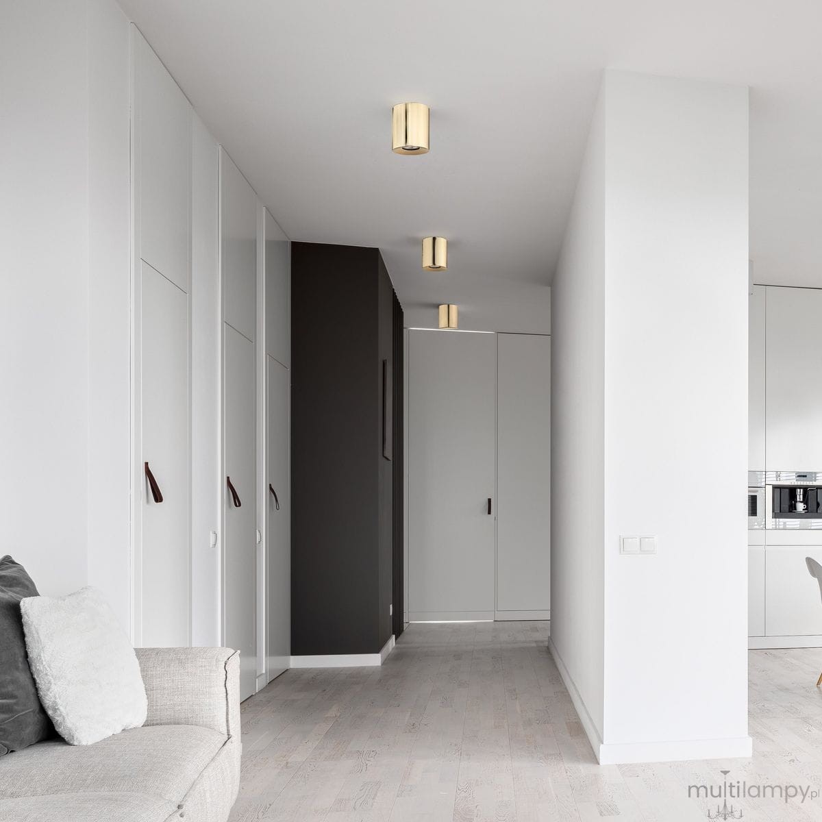 Złota lampa sufitowa Orion 1xGU10, cylindryczny kształt, zamontowana na białym suficie. Widoczna w nowoczesnym wnętrzu z szafkami w odcieniach bieli i szarości, w tle ciemna ściana. Oświetlenie w górnej części zdjęcia.