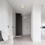Złota lampa sufitowa Orion 1xGU10, cylindryczny kształt, zamontowana na białym suficie. Widoczna w nowoczesnym wnętrzu z szafkami w odcieniach bieli i szarości, w tle ciemna ściana. Oświetlenie w górnej części zdjęcia.